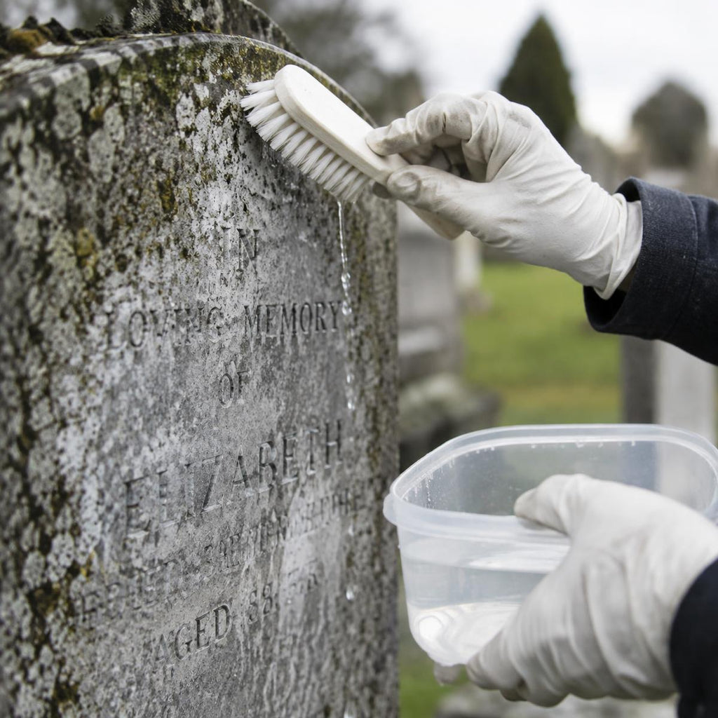 A close-up of gloved hands cleaning an engraved stone gravestone with a brush and holding a small plastic container of water in an outdoor cemetery setting.