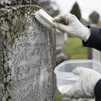 A close-up of gloved hands cleaning an engraved stone gravestone with a brush and holding a small plastic container of water in an outdoor cemetery setting.