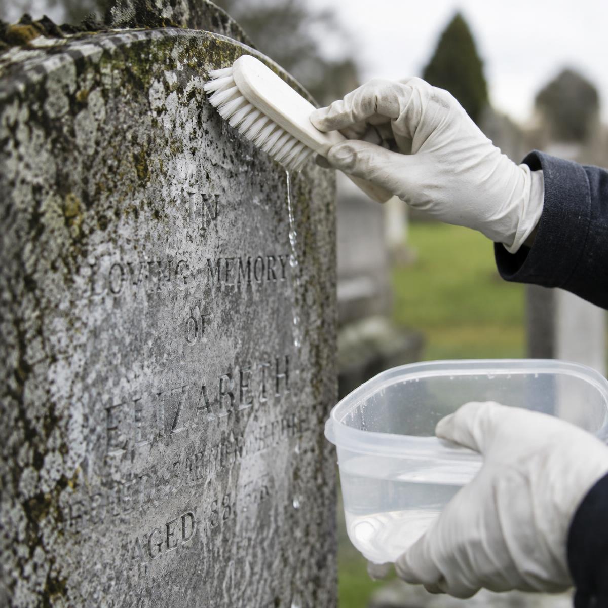 A close-up of gloved hands cleaning an engraved stone gravestone with a brush and holding a small plastic container of water in an outdoor cemetery setting.