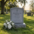 A gray stone gravestone in a grassy cemetery with trees and other headstones in the background. A white floral arrangement rests at the base of the gravestone, which is engraved with the name Jonathan M.