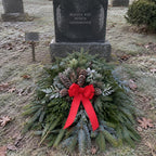 A festive evergreen wreath with pinecones and a large red ribbon bow placed in front of a gravestone on a grassy, lightly frosty outdoor cemetery ground.