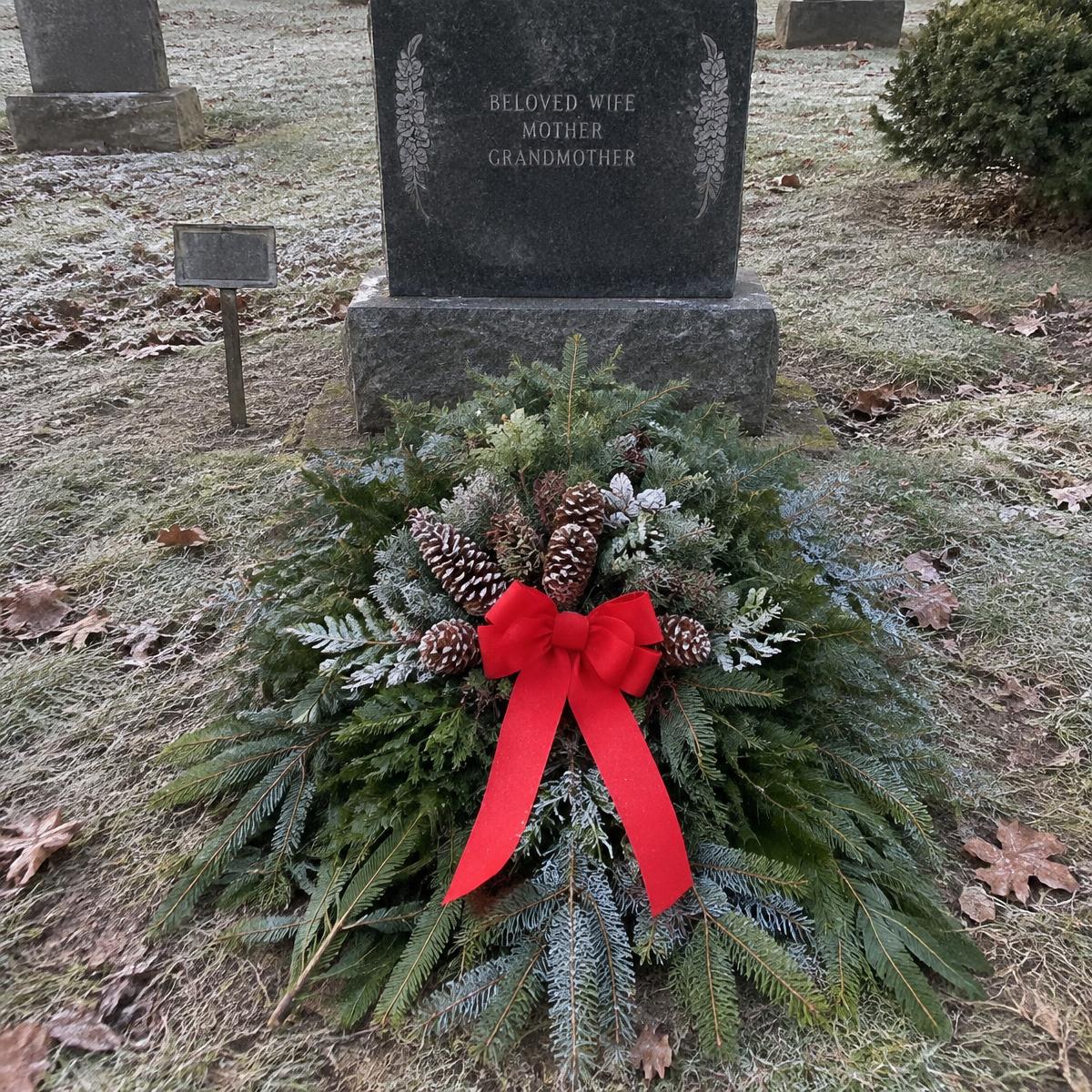 A festive evergreen wreath with pinecones and a large red ribbon bow placed in front of a gravestone on a grassy, lightly frosty outdoor cemetery ground.