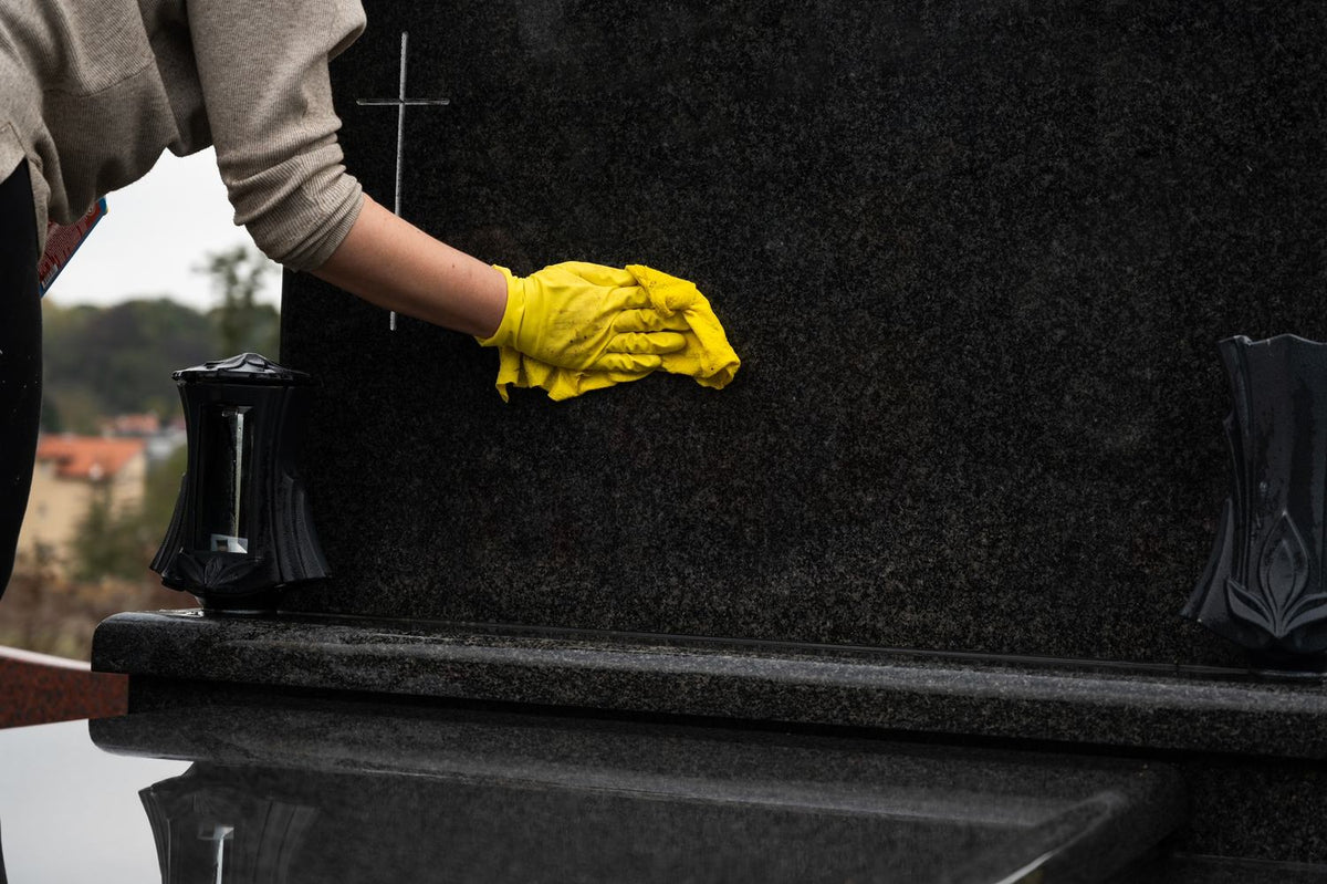 A person wearing a gray long-sleeve shirt and a bright yellow glove wipes a dark gray granite tombstone. A black metal casket is visible in the foreground, and decorative black vases sit on either side of the tombstone.